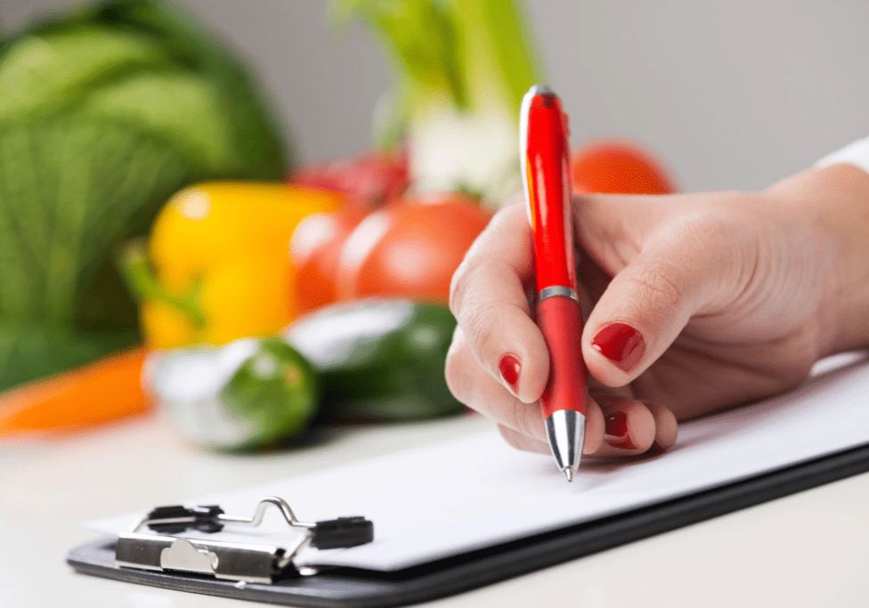 A person writing on paper with vegetables in the background.